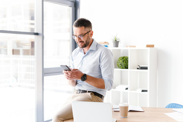 Image of modern business man in formal wear sitting on table in office near big window, and using...