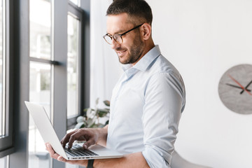 Image of satisfied office man wearing white shirt expressing success, while holding silver laptop during work in business interior