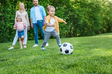 family looking at little boy playing with soccer ball in park