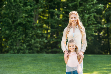 beautiful happy mother and daughter standing together and smiling at camera in park