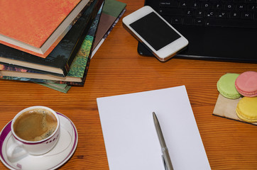 White paper with a pen, books, telephone, macarons and coffee on a wooden table