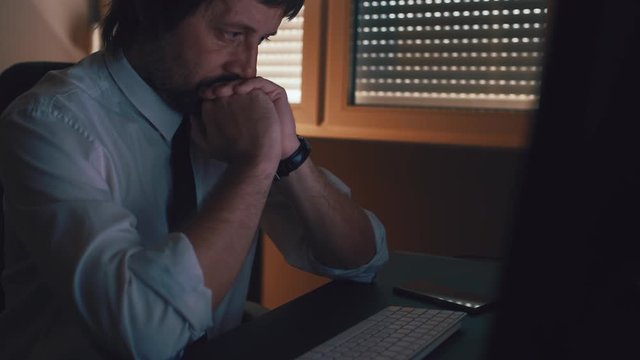 Close up face portrait of concentrated businessman thinking with hands clasped in front of him in dark office interior. Worried and concerned male business person looking at computer monitor