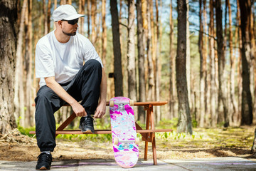 man in a white cap with a skateboard sits on a picnic table and tie a shoe in the park © superelaks