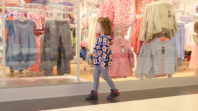 Kid shopping - little girl walks along glass clothes shop frontage with child mannequin