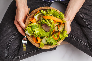 The woman in black jeans eating bright summer vegetable salad in a wooden bowl. Healthy vegan food concept.