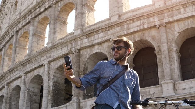 Young hipster man with bike at colosseum taking selfies pictures with smartphone in Rome city centre on sunny day slow motion steadycam