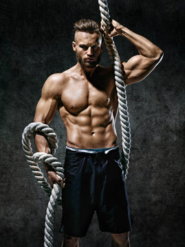 Athletic strong man with heavy ropes in hands. Photo of young man with naked torso on dark background. Strength and motivation