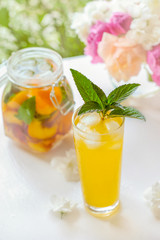 Pitcher with lid with water, detox with pieces of peach and apricot, with ice. A refreshing drink on a green background with a yellow rose.