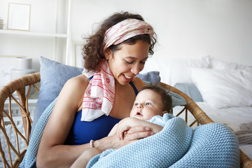 Portrait of cheerful joyful young Hispanic mother relaxing in weaven chair at home, talking to her...