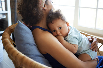 Rear view of young dark skinned female sitting by window in chair, nursing nine month old toddler....