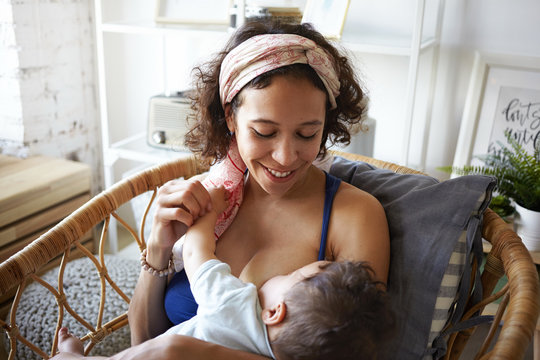 Horizontal shot of happy young mixed race mother with curly hair enjoying intimate moment with her baby son, sitting in weaven chair in bedroom, breastfeeding him, having joyful facial expression