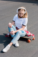 stylish teen girl listening music in headphones while sitting on skateboard