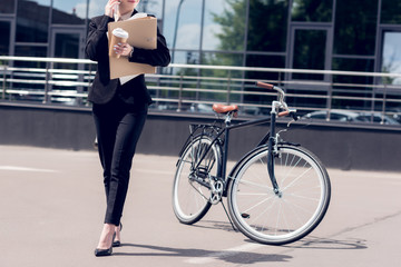 Fototapeta na wymiar cropped shot of businesswoman with documents and coffee to go talking on smartphone while walking on street with bicycle parked behind
