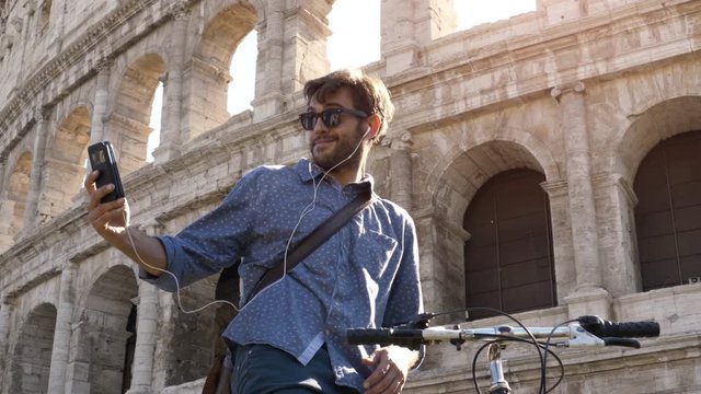 Young hipster man with bike at colosseum taking selfies pictures with smartphone in Rome city centre on sunny day slow motion steadycam