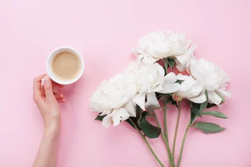 Tuinposter Woman hand with cup of coffee and beautiful white peony flowers on pink pastel table top view. Cozy breakfast in flat lay style. © juliasudnitskaya