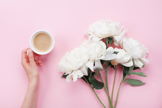 Woman hand with cup of coffee and beautiful white peony flowers on pink pastel table top view. Cozy breakfast in flat lay style.