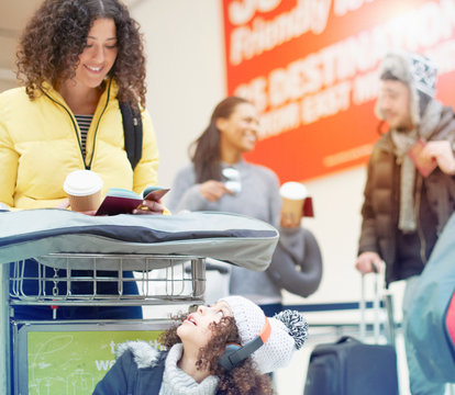 Girl looking up at mother looking at passport on luggage trolley in airport departure lounge
