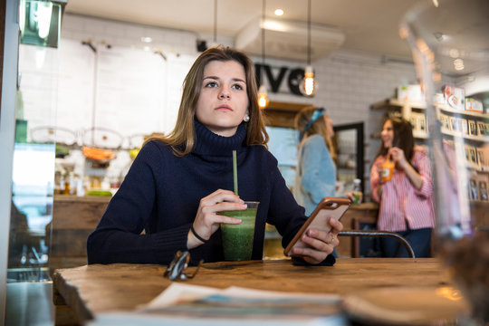 Young woman sitting in cafe, holding smartphone, drinking smoothie