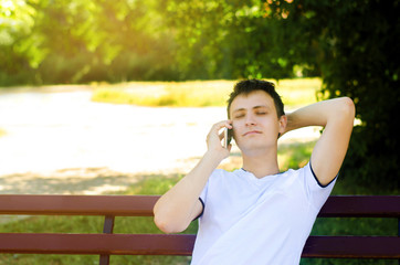 A young european guy is sitting on a bench in the park and talking on the phone, throwing his arm behind his head and closing his eyes. Relaxed phone conversation, fatigue, tranquility, anti-stress.