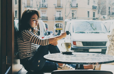Charming asian female taking selfie by a camera of a mobile phone while sitting in a modern city coffee shop with big windows. Hipster girl having video call while drinking coffee or tea in a cafe.