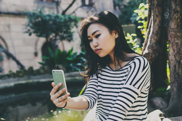 Beautiful model look asian hipster girl with long dark hair in a striped longsleeve t-shirt is taking selfie by a camera of the mobile phone while sitting on a natural plants background on a sunny day