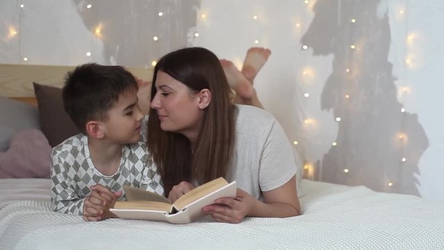 Beautiful girl with long hair reads a book to a small boy lying on a bed and kisses him