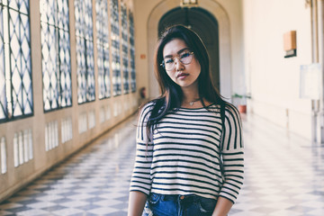 Stylish asian woman wearing glasses, striped long sleeve t-shirt and blue jeans walking the museum corridor on a day off.  Hipster girl visiting new trendy art space on a spring day.