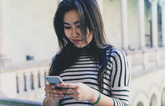 Photo of a beautiful model look asian female with long black hair looking at the smartphone display. Travel blogger wearing casual clothes reading email on a mobile phone connected to wifi.