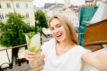 Let's drink. Closeup of emotional posistive woman doing selfie photo and clinking lemonade glasses with camera and smiling while standing on terrace of modern city cafe. European city view behind her.