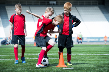 Full length portrait of junior football team practicing in stadium, focus on boy leading ball round...