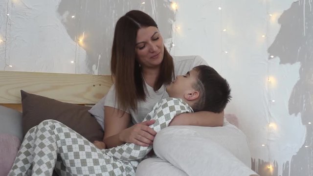A beautiful mother plays with her son sitting on the bed and kisses him having a good mood.