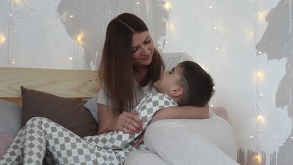 A beautiful mother plays with her son sitting on the bed and kisses him having a good mood. Overall plan