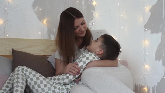 A beautiful mother plays with her son sitting on the bed and kisses him having a good mood. Overall plan