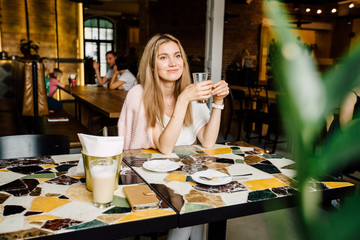 Young blond lady drinking coffe and looking aside through a window. in modern cafe interior.