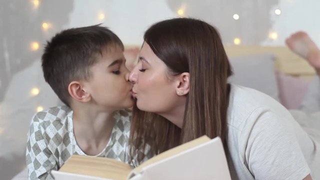 A beautiful girl with long hair reads a book to a small boy lying on a bed and kisses him.HD