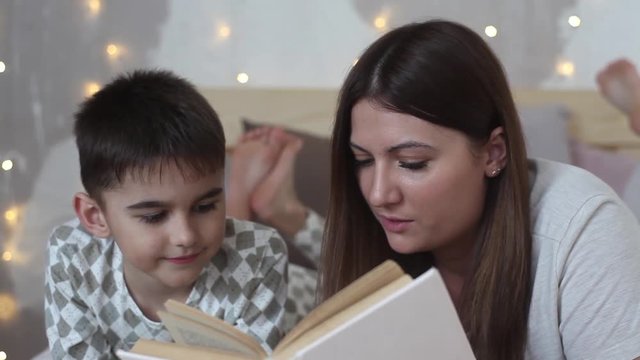 A beautiful mother with long hair reads a book to a small boy lying on a bed and kisses him having a good mood.HD