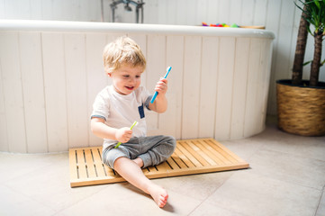 A toddler boy with a toothbrush sitting on the floor in the bathroom at home.