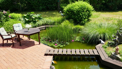 attraktiver naturbelassener Teich an rote Terrasse angrenzend mit Brücke und Tisch und Stühlen