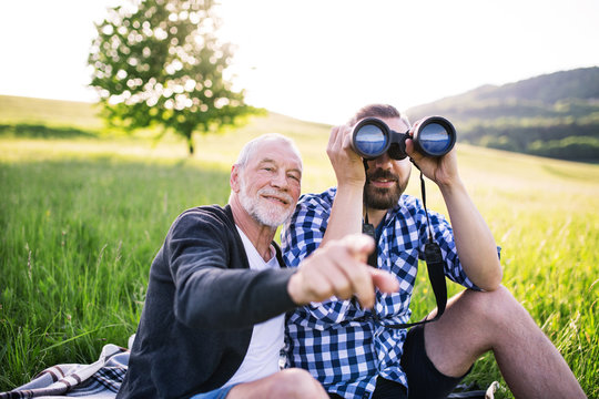 An adult hipster son with binoculars and senior father sitting on the grass in sunny nature.