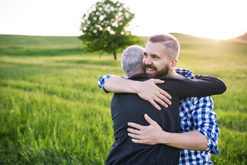An adult hipster son with senior father on a walk in nature at sunset, hugging.