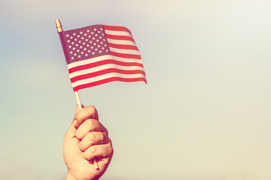 Woman hand holding american flag of the USA with stars and stripes against blue sky. Vintage toned effect.