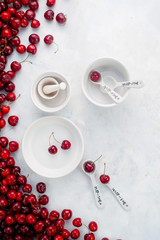 Cherries and white porcelain baking dishes with measuring spoons on a white stone background. Minimalist cherry pie baking concept.
