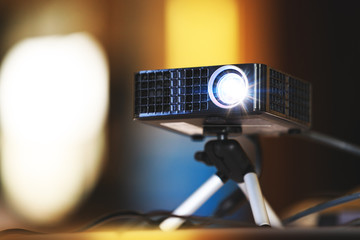 Real projector at business conference or presentation in the office room. Close up of projector for cinema and video projections. Black projector on tripod in hall or seminar meeting room.  - obrazy, fototapety, plakaty