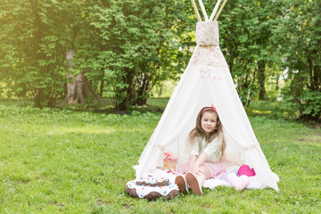 Little girlplaying in a tent, children's house wigwam in park. Happy mothers day. Child with big donuts and straw hat.  Summer, happy childhood concept. Cute emotions girl with pink bow. © MartaKlos