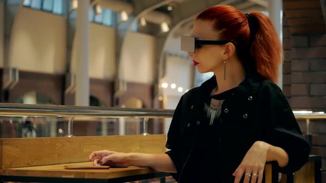 Stylish red hair woman with square sunglasses watches mobile phone in the mall