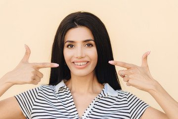 Portrait of pleasant looking brunette female has appealing look, indicates with both fore fingers...