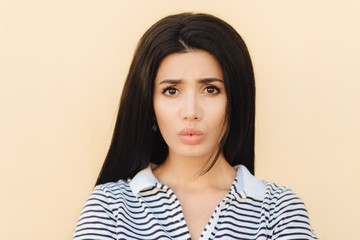 Fototapeta premium Gloomy brunette young female pouts lips in discontent, being dissatisfied with something, has healthy pure skin, dark eyes and make up, dressed in casual clothes, isolated over studio background