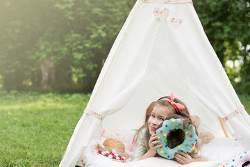 Little girl lying and playing in a tent, children's house wigwam in park. Happy mothers day. Child with big donuts and straw hat. Summer, happy childhood concept. Cute emotions girl with pink bow. © MartaKlos