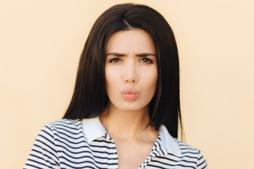 People, beauty and facial expressions concept. Discontent brunette cute woman pouts lips and frowns face, has sullen look, dressed in casual striped t shirt, has gloomy expression after quarrel