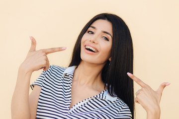 Portrait of happy gorgeous female model indicates with fore fingers at her face, advertises braces for even teeth, has dark long straight hair, shining eyes and perfect healthy skin, models indoor